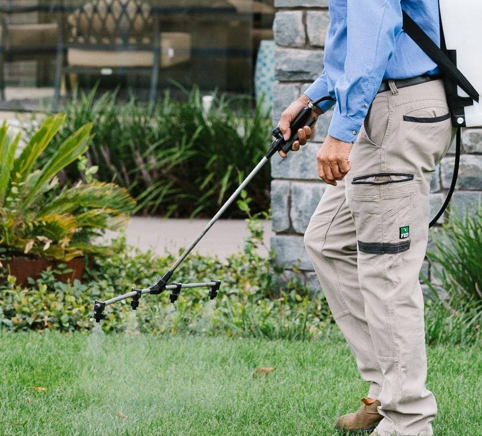 Technician spraying a home lawn beside a garden bed with a backpack sprayer and boom wand