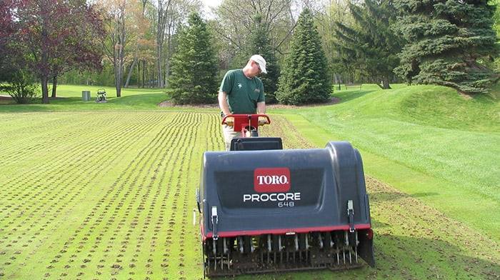 Operator coring green turf with a walk-behind aerator, leaving neat rows of holes