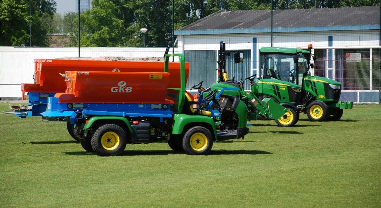 Top dressing spreaders on utility vehicles parked beside compact tractors on a sports field