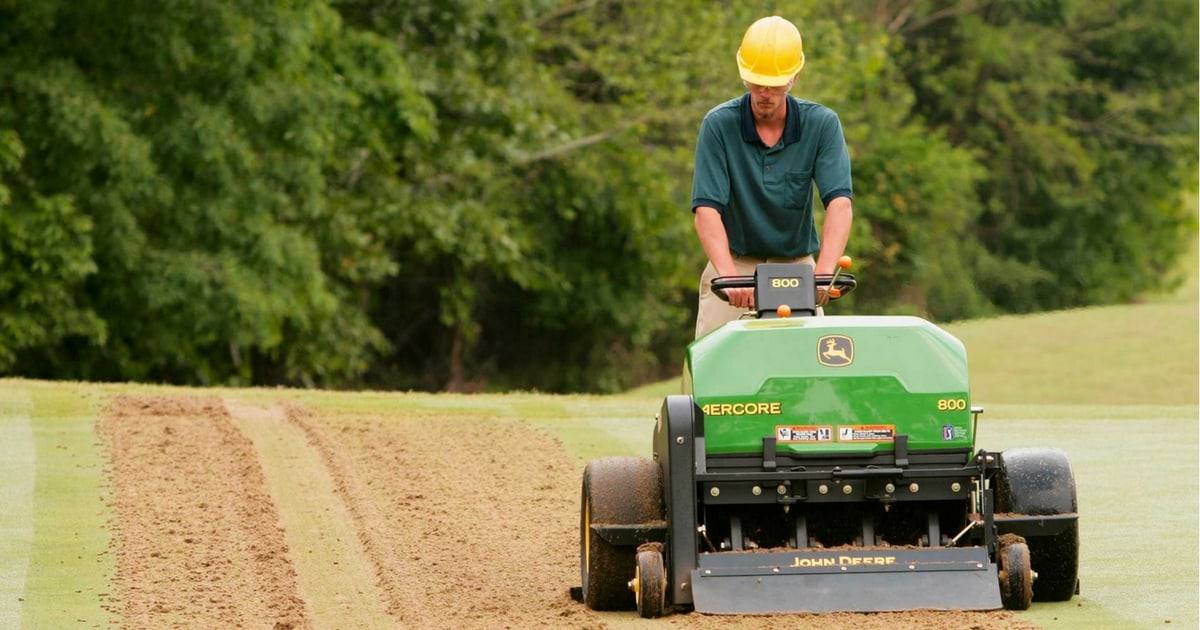 Operator running a walk-behind aerator across turf, leaving rows of soil cores behind